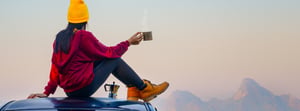 image of woman gazing out over far off mountains during fall with a hot cup of coffee while sitting on the roof of her car.